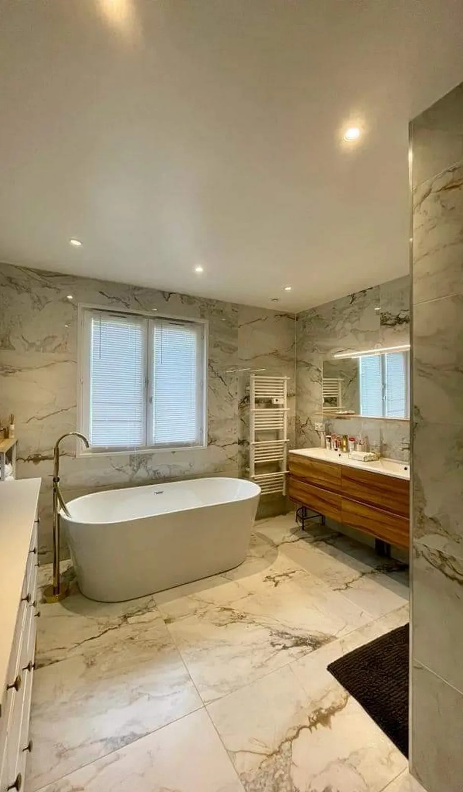Salle de bains terminee entierement revetue de carrelage grand format effet marbre veine beige et gris, avec baignoire ilot blanche, robinetterie sur pied doree, meuble vasque suspendu en bois et seche-serviettes