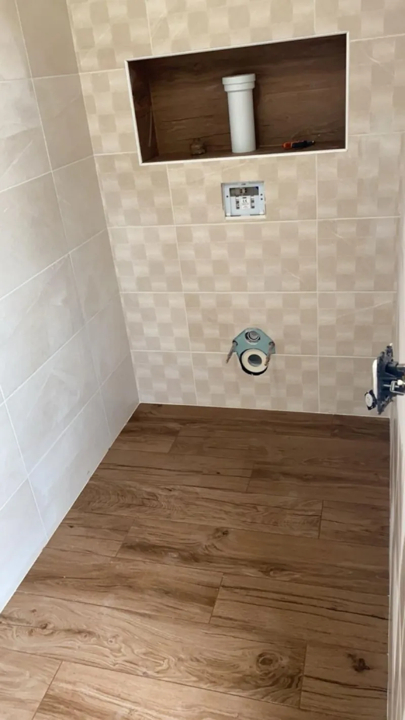 WC en cours de finition : murs en faience beige a motif damier, niche horizontale habillee de carrelage effet bois fonce, bati-support et sortie d'evacuation apparents, sol en carrelage effet bois
