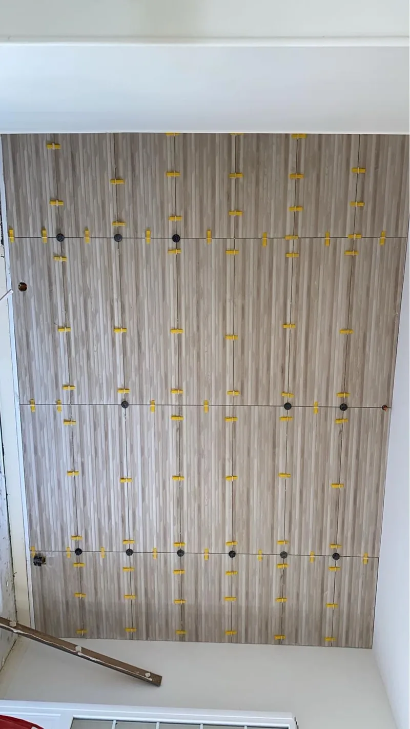 Mur habille de carrelage grand format a veinage strie vertical beige-gris, en cours de pose, maintenu par des croisillons autonivelants jaunes ; chape lissee claire au sol devant