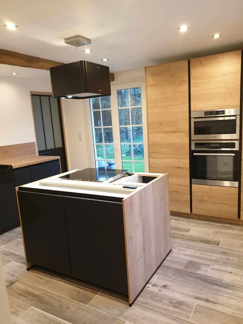 Cuisine ouverte avec sol carrele en grand format effet bois grise, ilot central a facades noires et panneau effet chene, hotte suspendue noire, colonne de fours