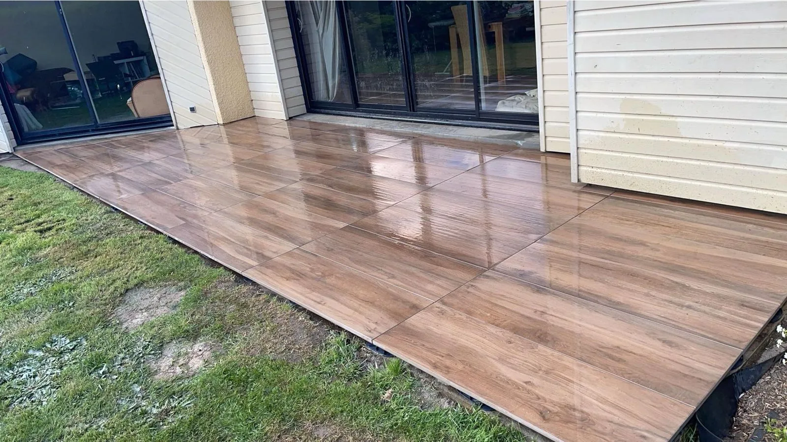 Terrasse exterieure en carrelage grand format effet bois, humide et reflechissante, longeant une maison a bardage creme avec grandes baies coulissantes, pelouse a gauche