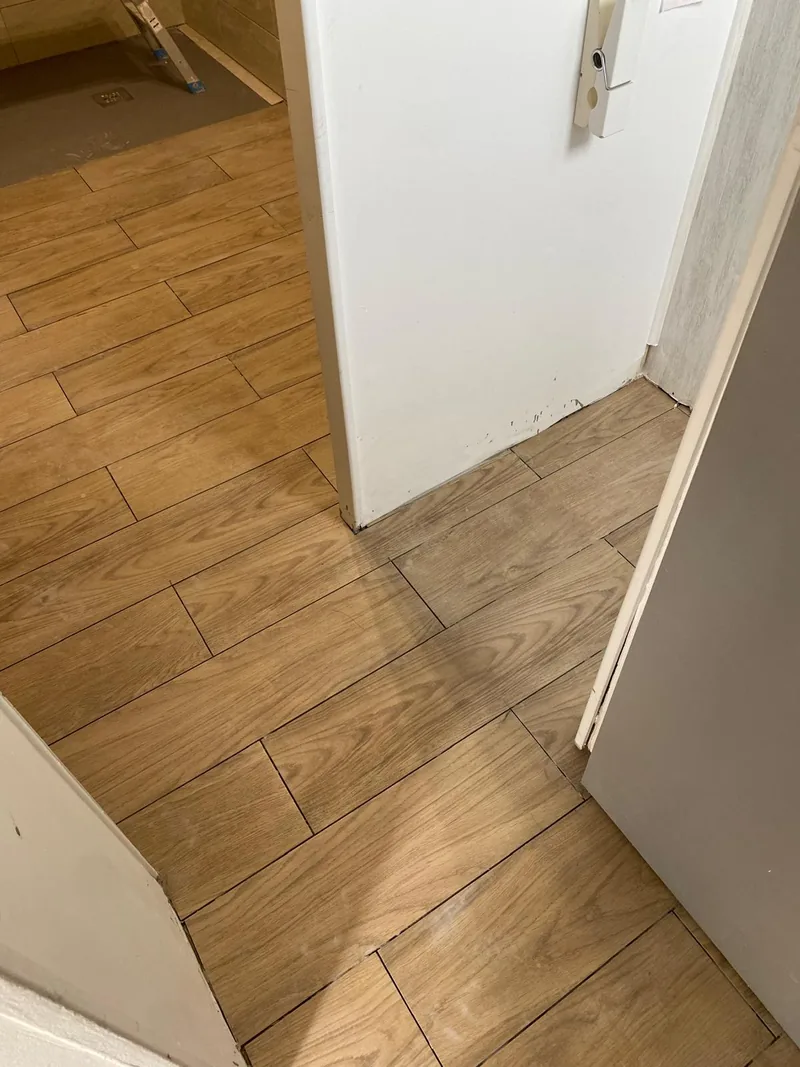 Passage de porte carrele en effet bois chene pose en decale, franchissant un seuil ; piece voisine encore en chape avec siphon de sol apparent