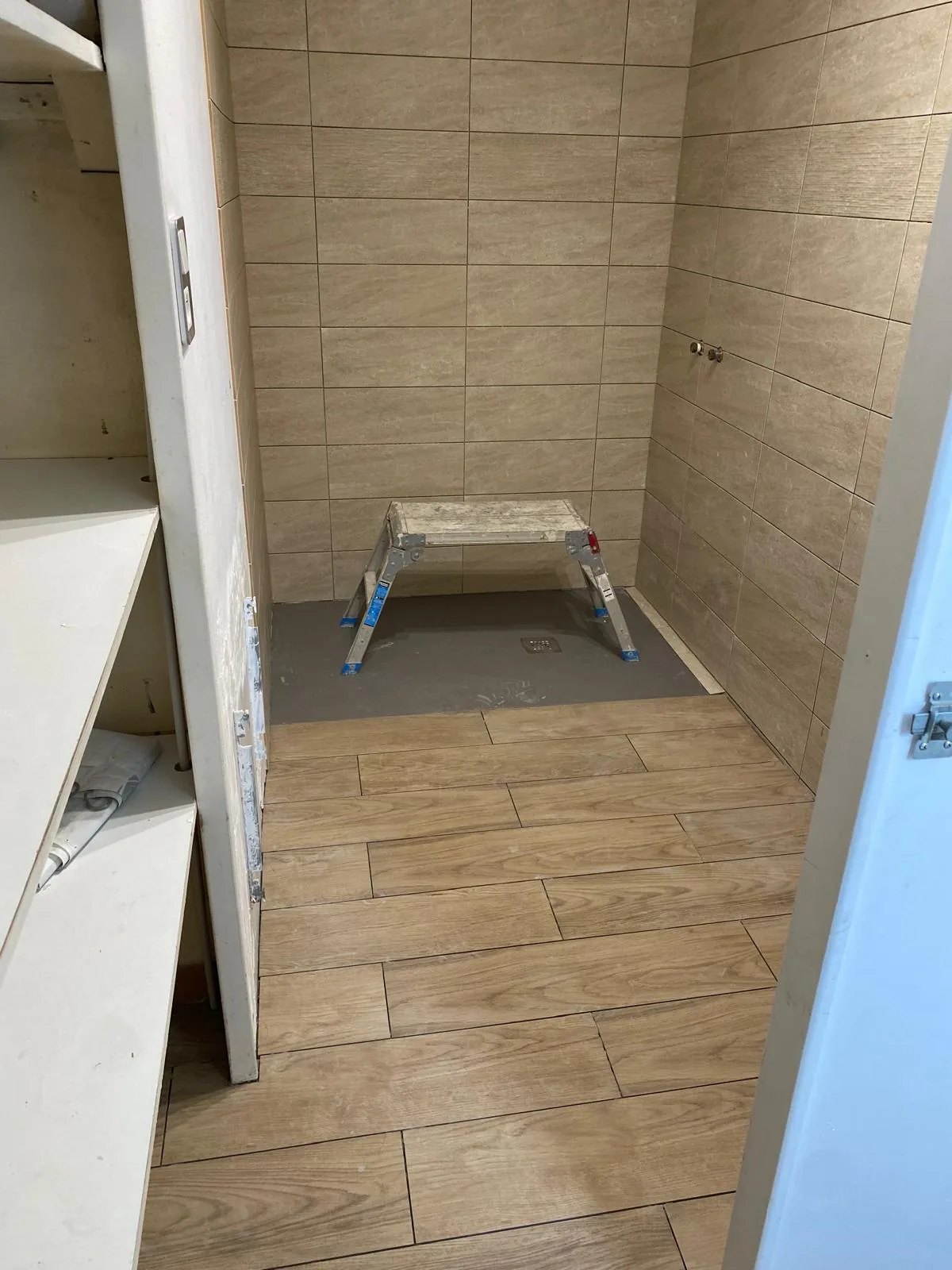 Salle de bains en cours de realisation : murs en faience effet travertin posee en decale, sol en carrelage effet bois, zone de douche encore en chape grise avec siphon de sol, marchepied pose au centre