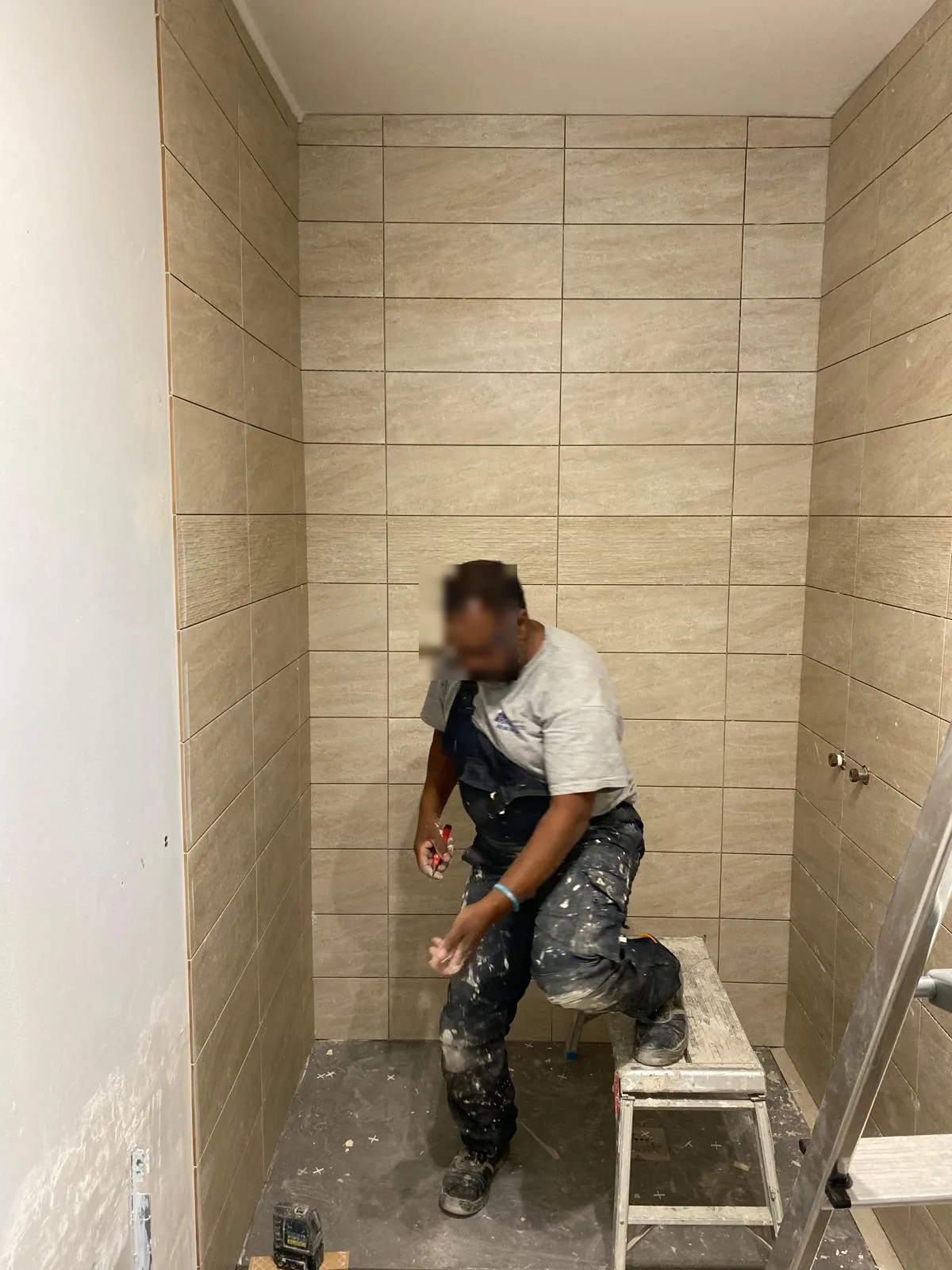 Carreleur au travail dans une niche de douche : murs en faience effet travertin posee en decale, sol encore en chape, marchepied et laser de chantier au sol