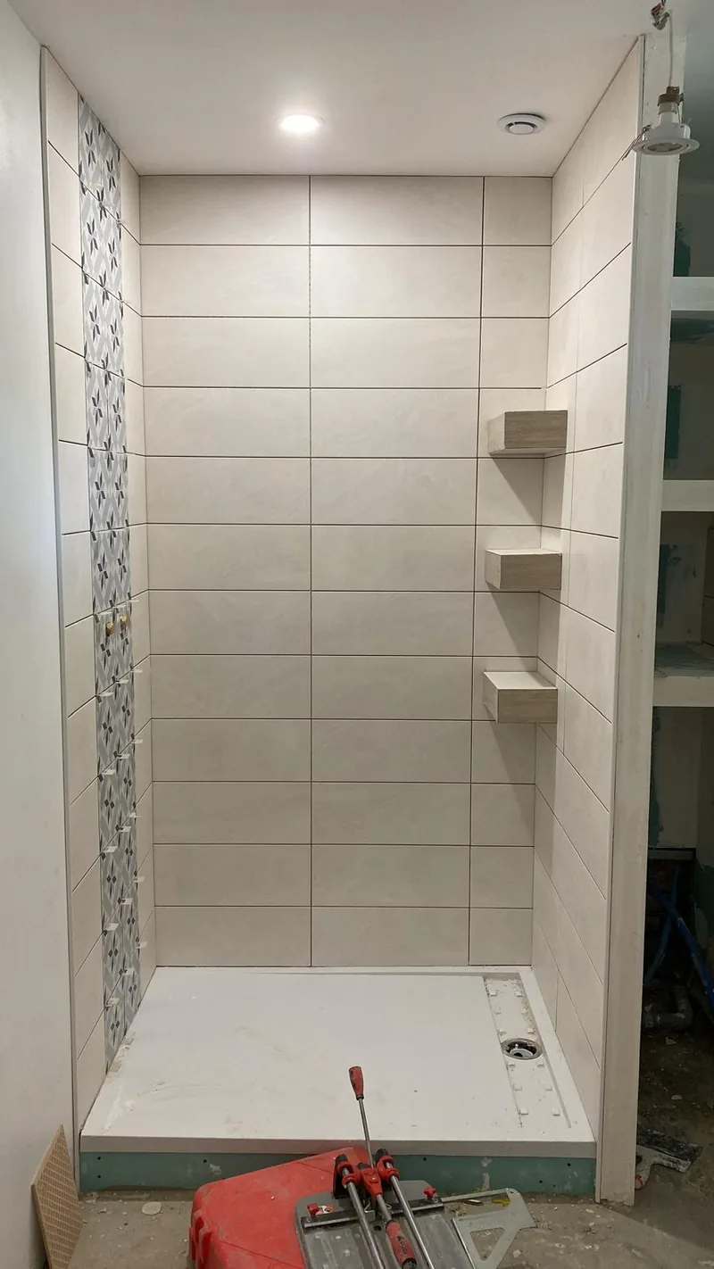 Douche en cours de realisation : murs en faience claire posee en decale, frise verticale a motif geometrique gris-bleu a gauche, trois etageres d'angle integrees a droite, receveur blanc, coupe-carreau pose au sol