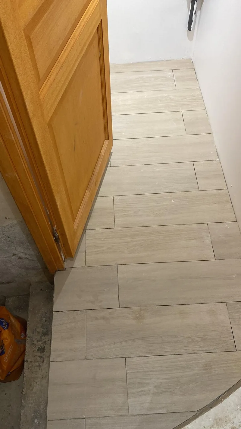 Sol carrele en effet bois beige clair pose en decale, le long d'une porte en bois verni ; bande de beton brut non carrelee a gauche