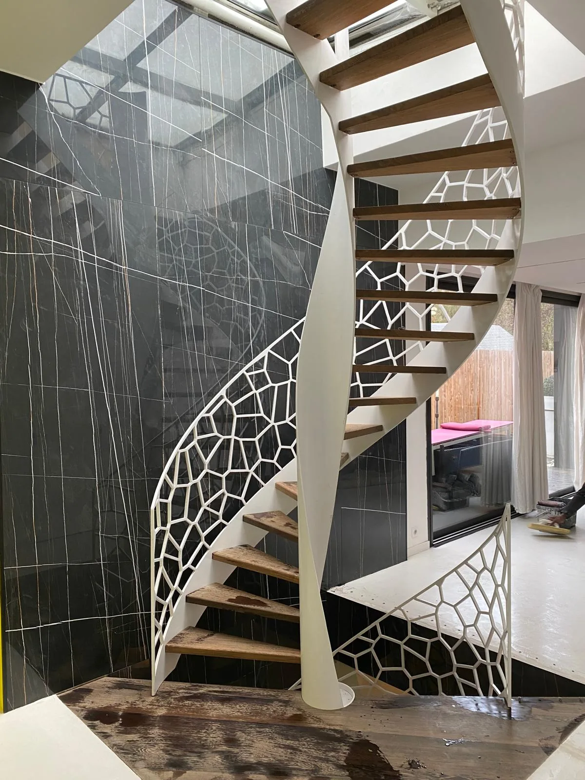 Mur double hauteur habille de dalles grand format effet marbre noir veine de blanc, autour d'un escalier helicoidal a limon blanc ajoure et marches en bois, sous une verriere