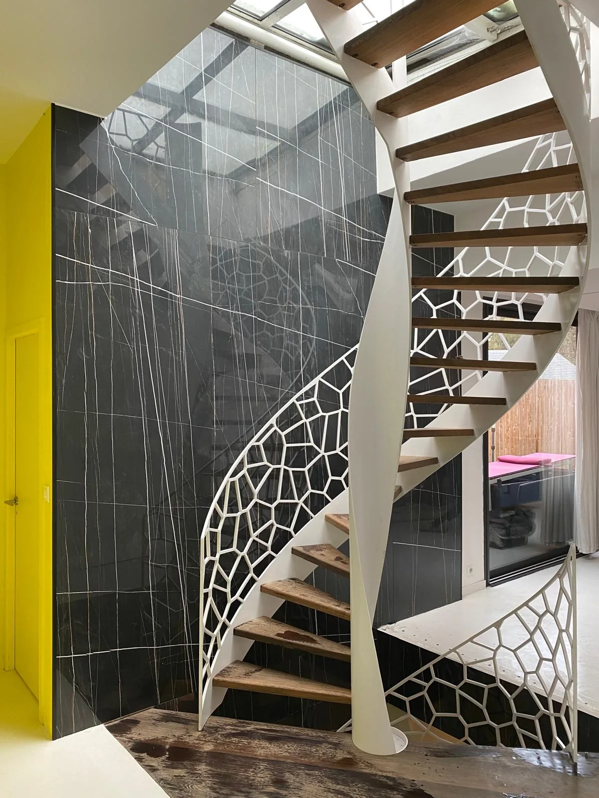Mur double hauteur habille de dalles grand format effet marbre noir veine de blanc, escalier helicoidal a limon blanc ajoure et marches en bois, mur jaune et verriere