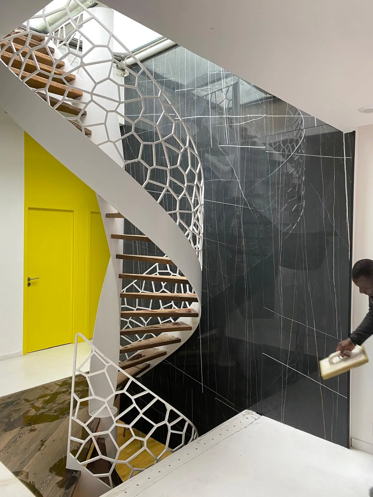 Mur interieur habille de grand format effet marbre noir veine blanc sur toute la hauteur, derriere un escalier helicoidal blanc a marches bois ; porte jaune sur la gauche