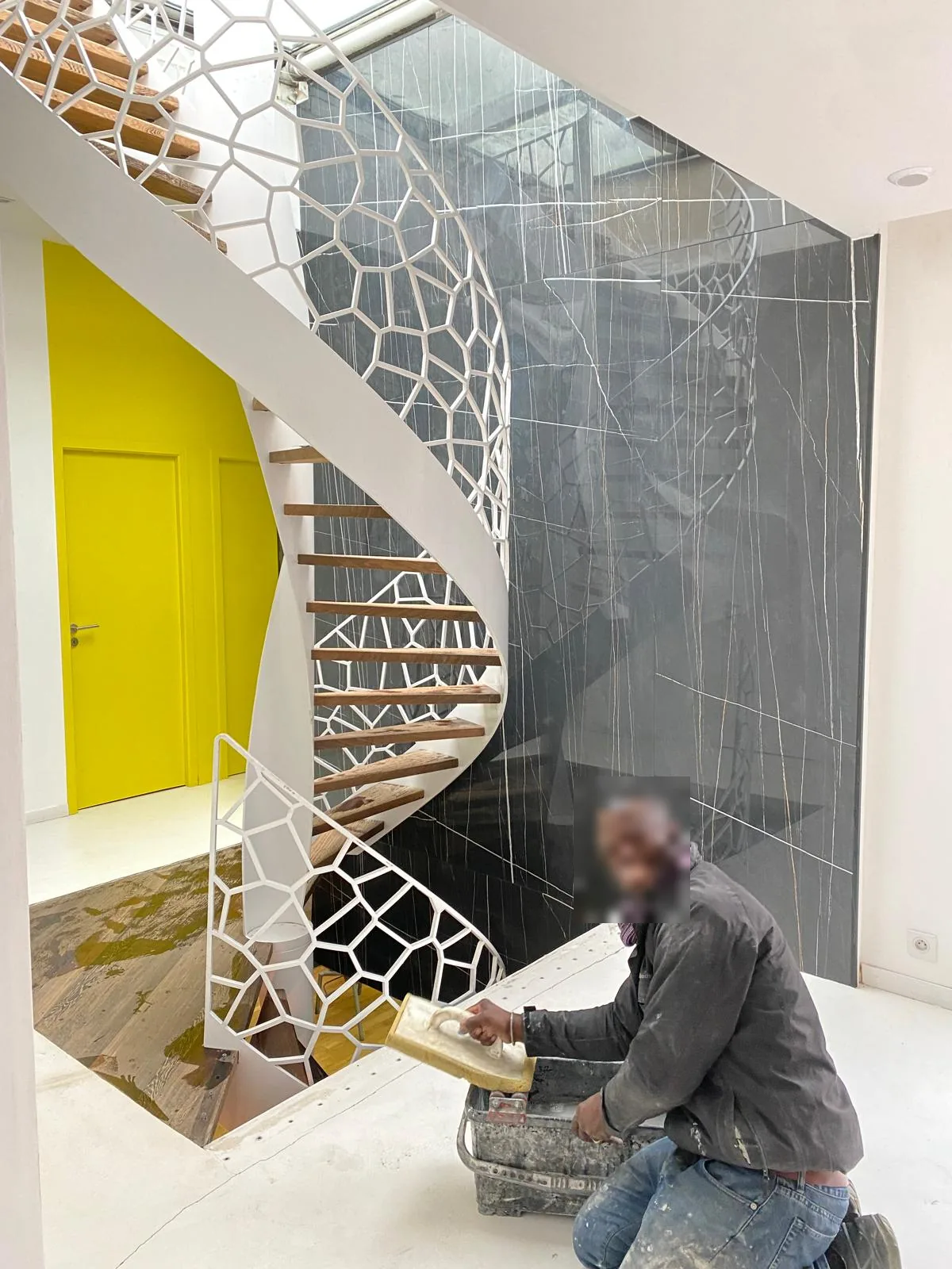 Homme au travail, taloche a la main, devant un mur interieur en grand format effet marbre noir veine blanc, au pied d'un escalier helicoidal blanc