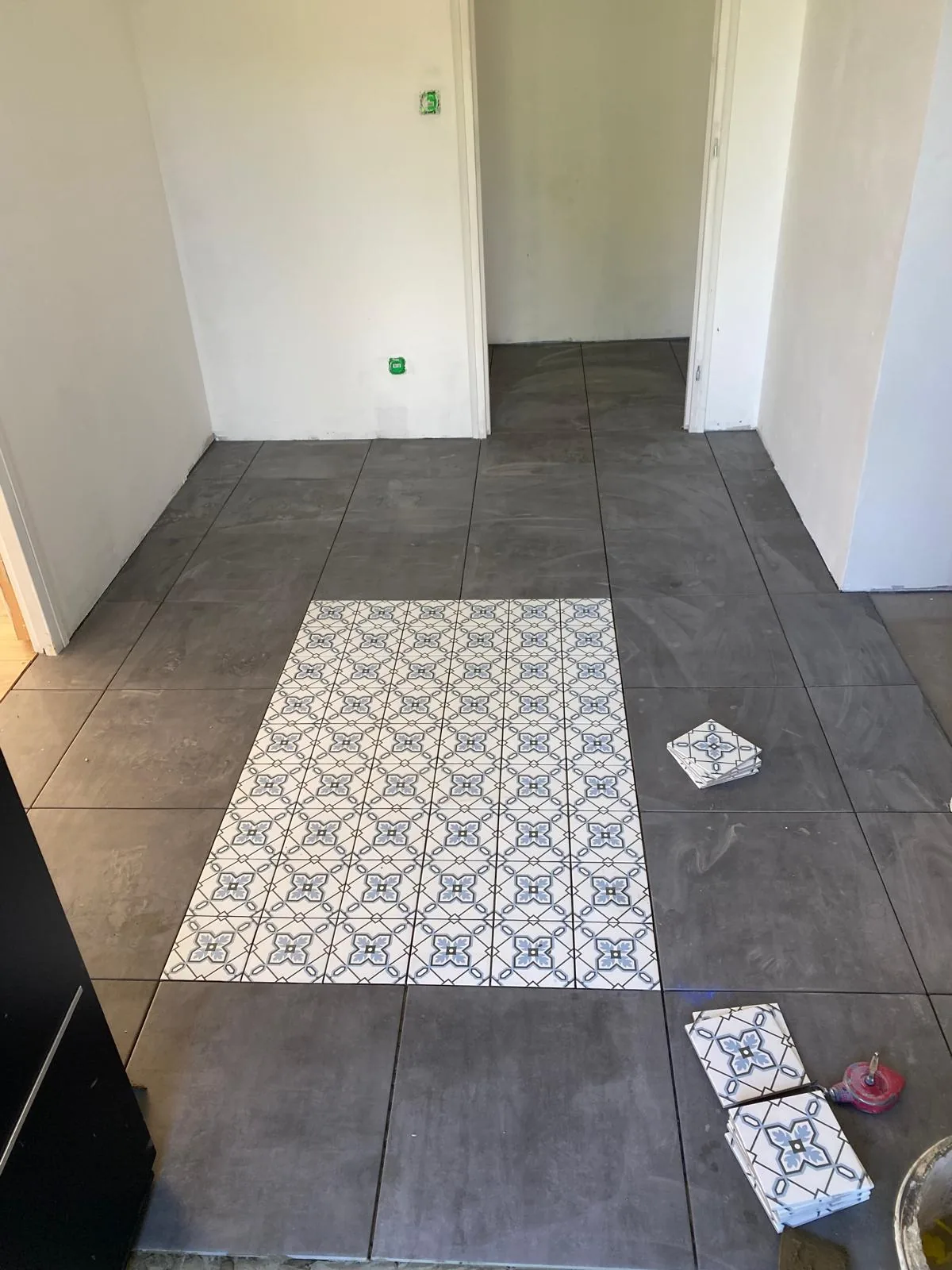 Sol carrele effet beton gris fonce avec un tapis rectangulaire de carreaux ciment a motifs bleus et blancs encastre au centre ; piles de carreaux et molette de coupe posees au sol