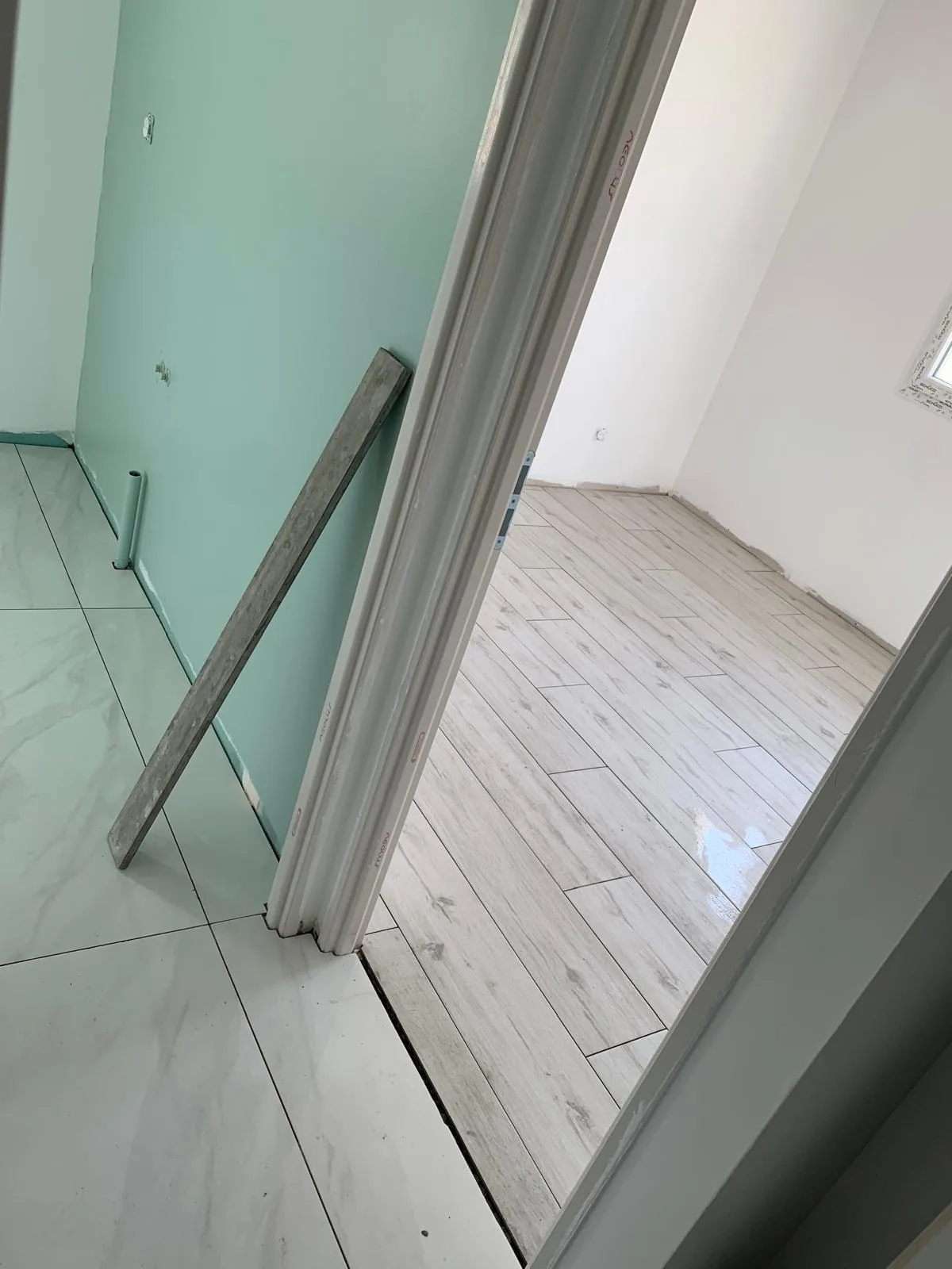Vue depuis un couloir carrele en effet marbre blanc vers une chambre au sol en carrelage imitation bois clair grise ; mur vert d'eau et bloc-porte non pose appuye au mur