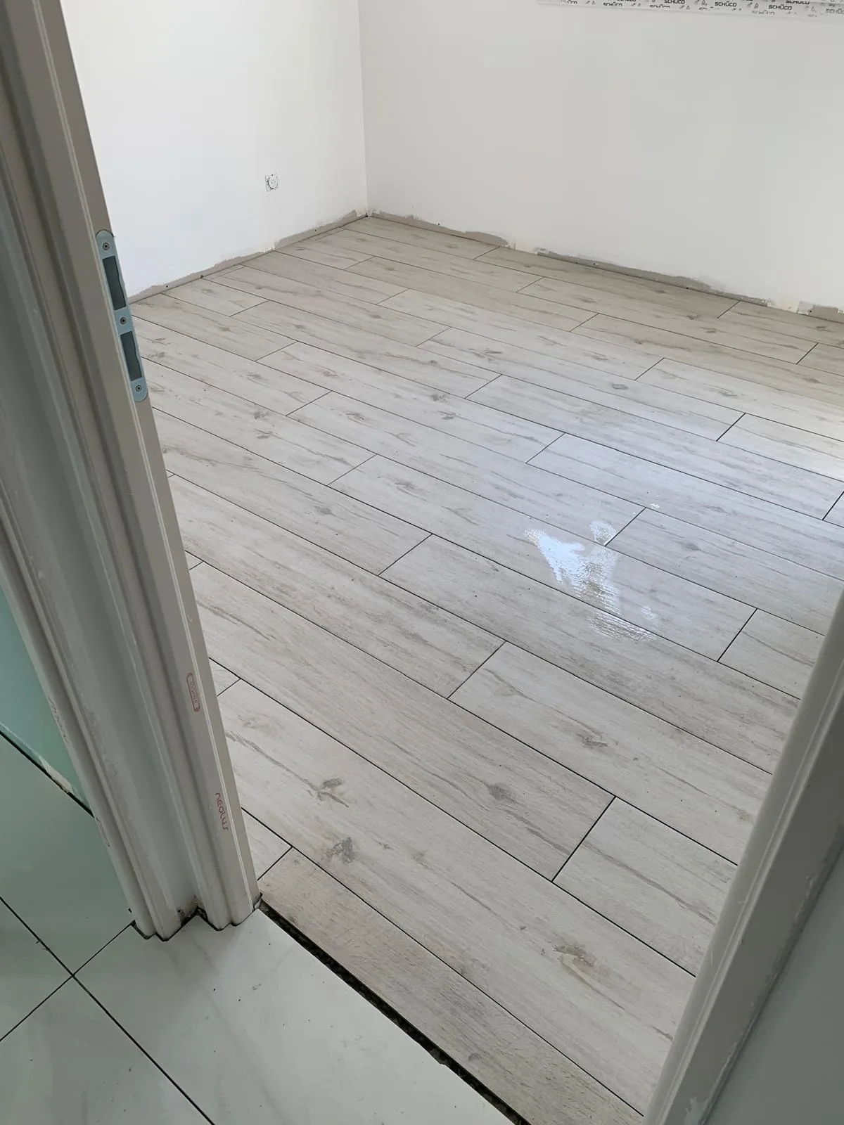Chambre au sol en carrelage imitation bois clair pose en lames a coupe decalee, avec la jonction visible vers le carrelage effet marbre blanc du couloir