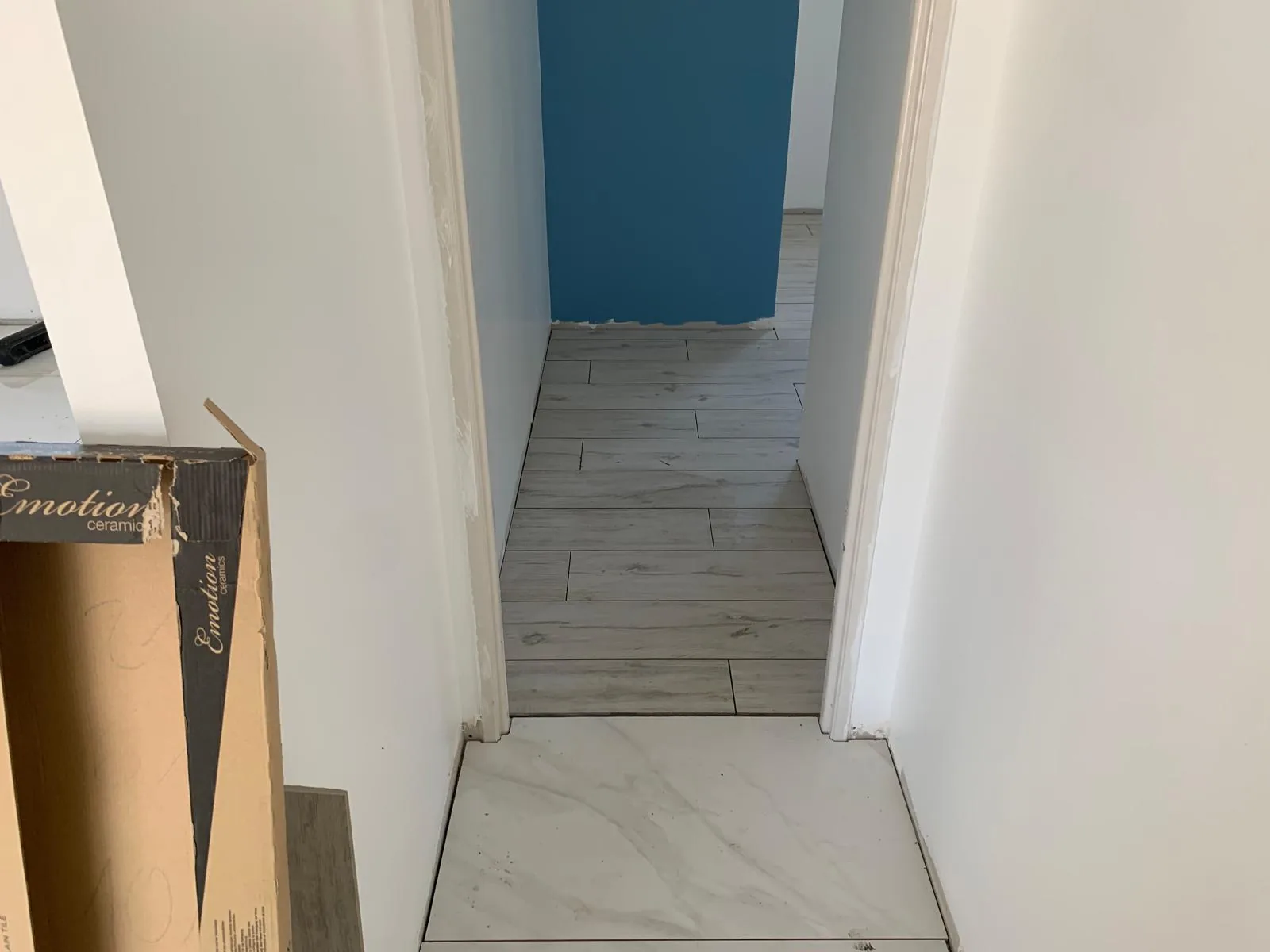 Couloir etroit au sol en carrelage imitation bois clair, prolonge par un degagement en carrelage effet marbre blanc ; mur bleu au fond