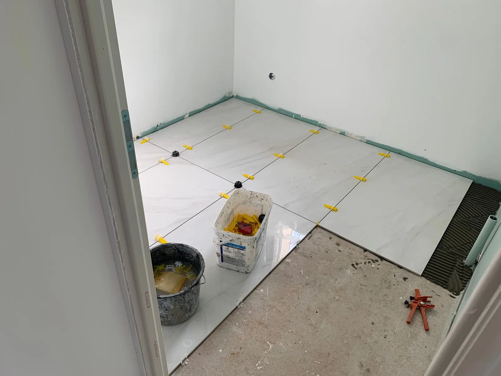 Petite piece au sol en cours de carrelage grand format effet marbre blanc, croisillons autonivelants jaunes sur les joints, colle peignee et pince a croisillons au sol