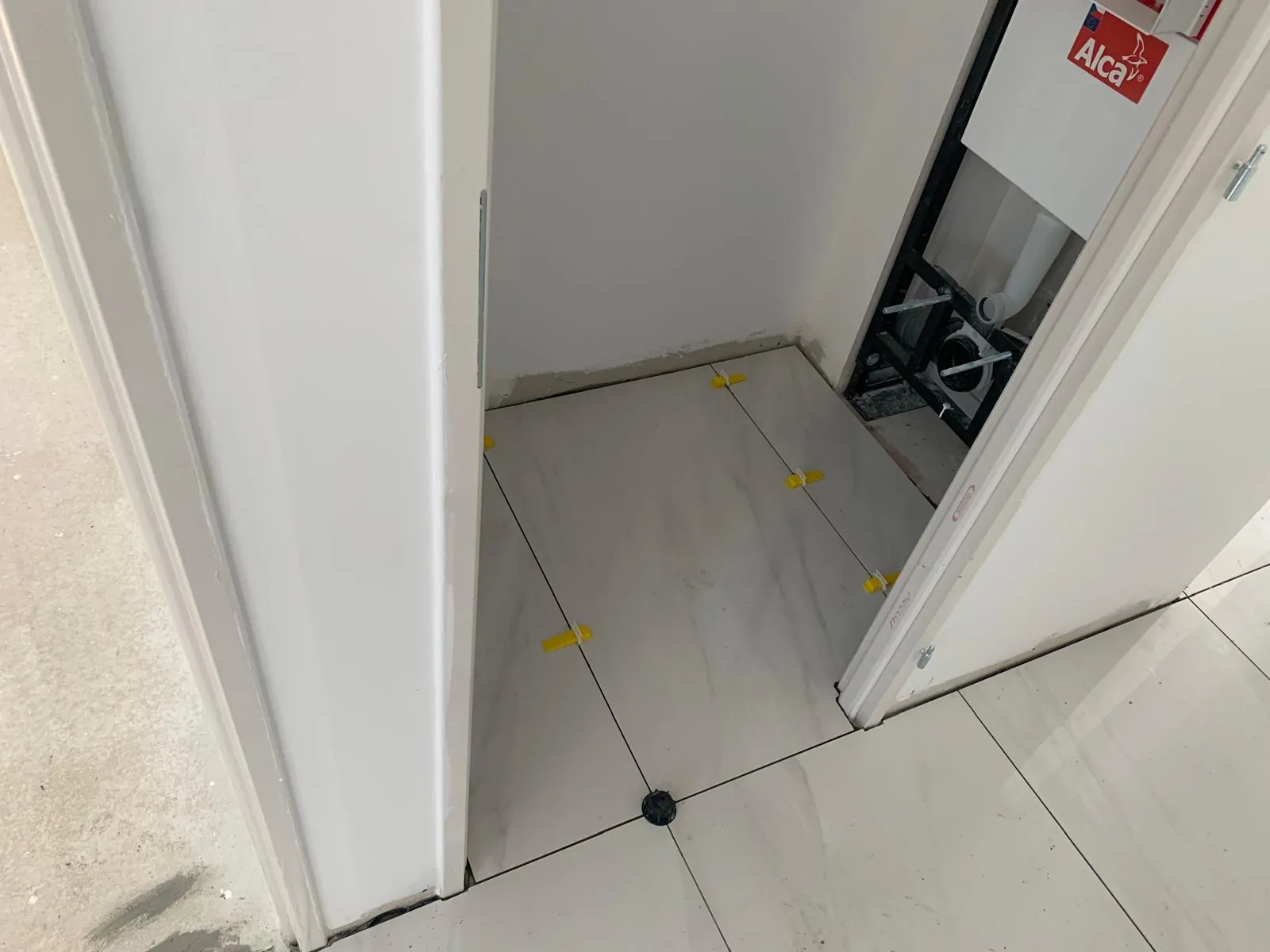 WC en chantier au sol carrele en grand format effet marbre clair avec croisillons autonivelants jaunes ; bati-support de WC suspendu et son reservoir encastre visibles a droite