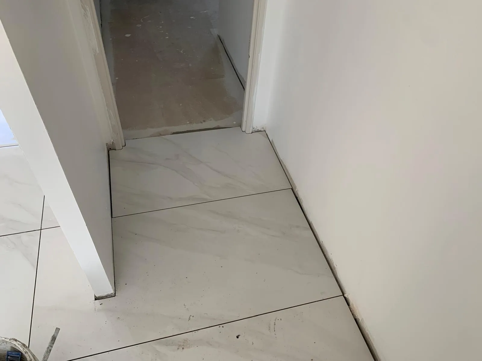 Sol en carrelage grand format effet marbre blanc posé jusqu'au seuil d'une porte, la pièce attenante restant en chape brute