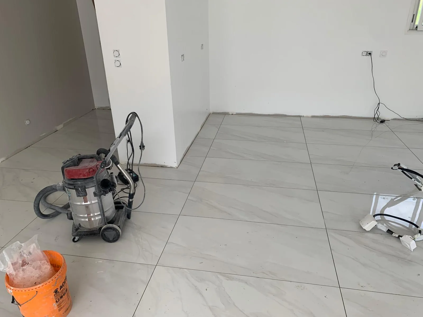 Pièce en chantier avec sol en carrelage grand format effet marbre blanc brillant, aspirateur de chantier et seau au premier plan