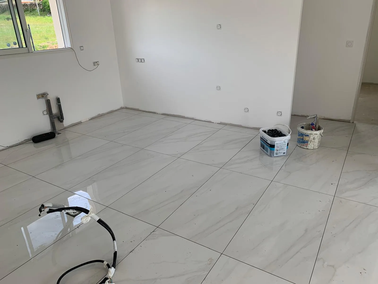 Sol en carrelage grand format effet marbre blanc dans une pièce aux murs blancs, avec une ouverture vitrée donnant sur de la verdure