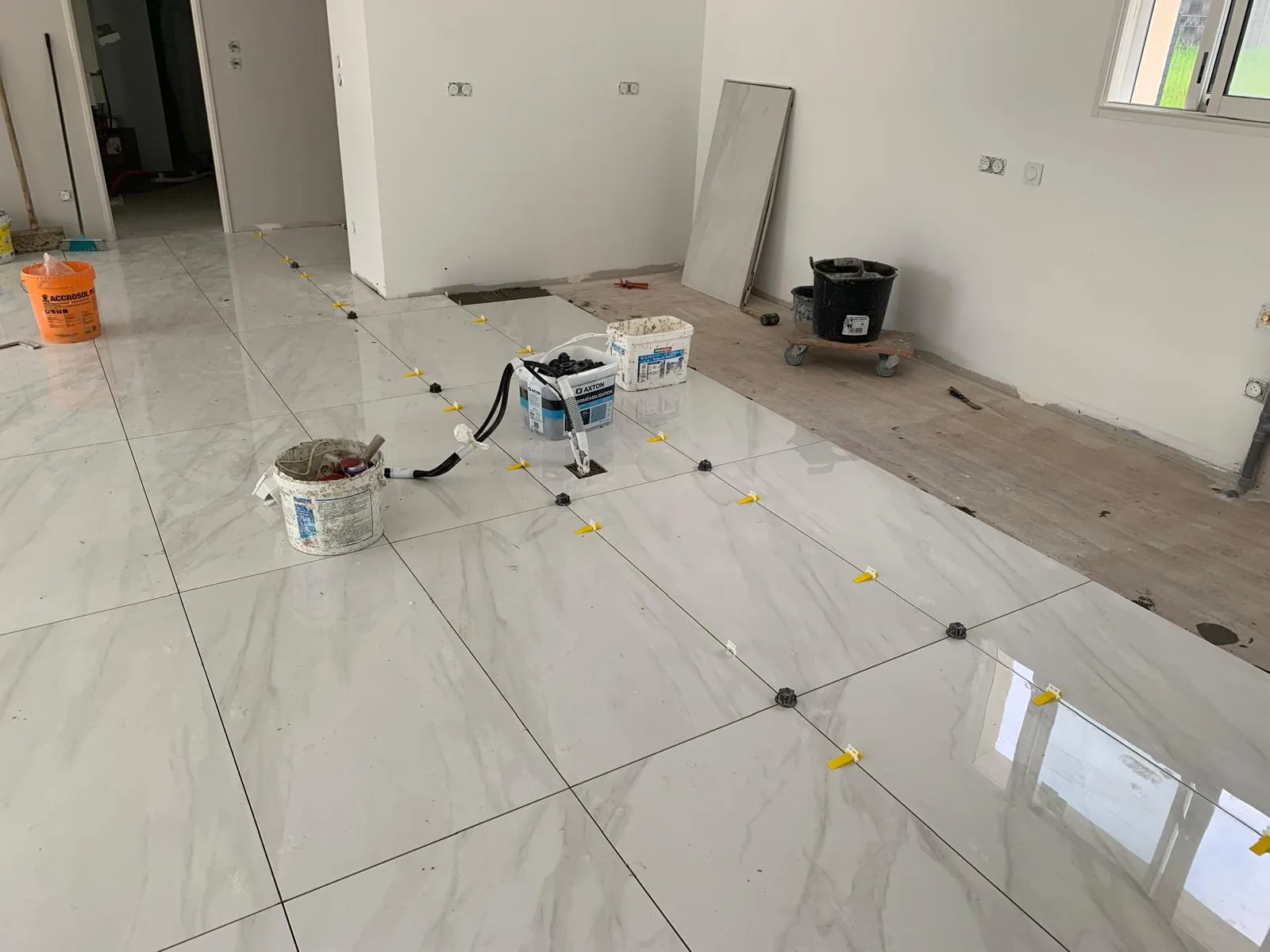 Sol en carrelage grand format effet marbre blanc en cours de pose, avec cales jaunes au sol et seaux de chantier