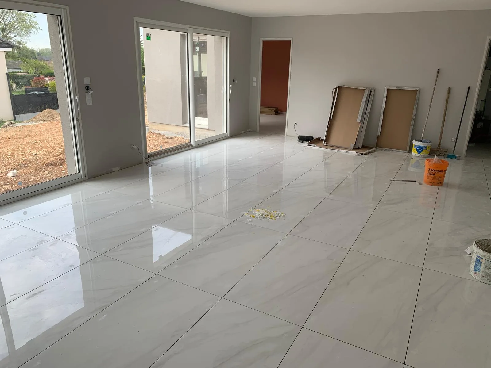 Vaste pièce à vivre en chantier avec un sol en carrelage grand format effet marbre blanc brillant très réfléchissant et deux baies vitrées donnant sur un terrain en cours d'aménagement