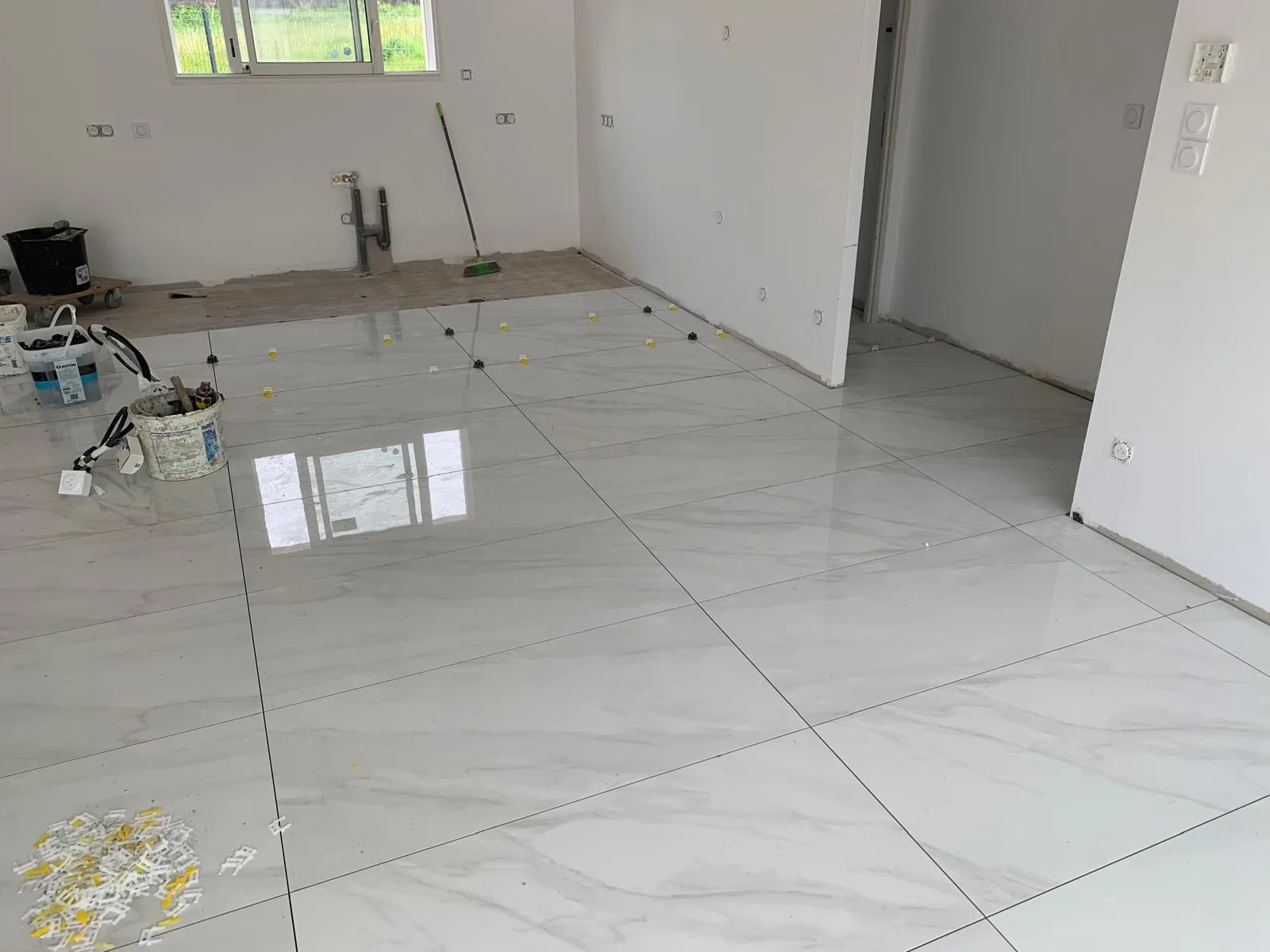 Pièce en chantier avec sol en carrelage grand format effet marbre blanc brillant, fenêtres sur la gauche et matériaux entreposés