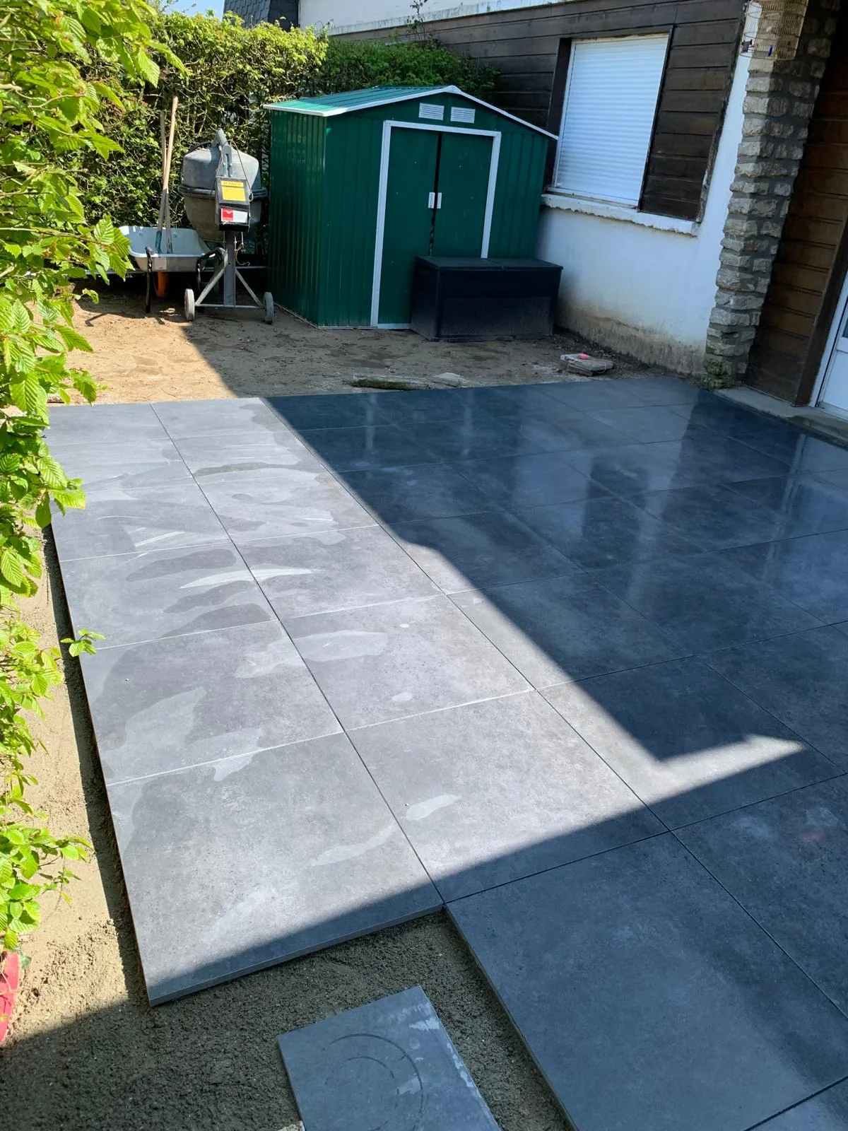 Terrasse extérieure en carrelage grand format gris en cours de pose au pied d'une maison, avec un abri de jardin vert et une bétonnière en arrière-plan