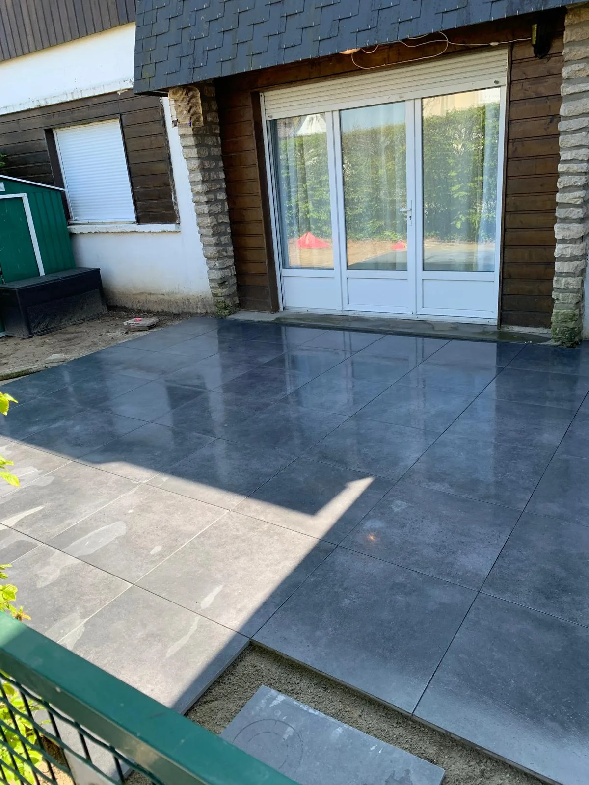 Terrasse extérieure en carrelage grand format gris posée le long d'une maison avec baie vitrée, bordée d'une haie