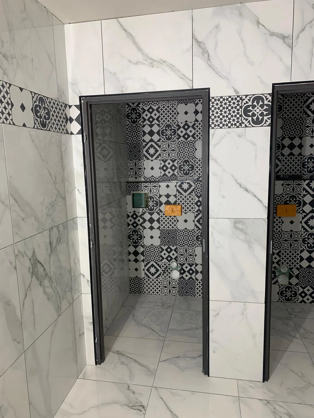 Deux cabines de douche côte à côte, murs en faïence grand format effet marbre blanc, fond et frise horizontale en carreaux à motifs noir et blanc, parois vitrées à profilés sombres