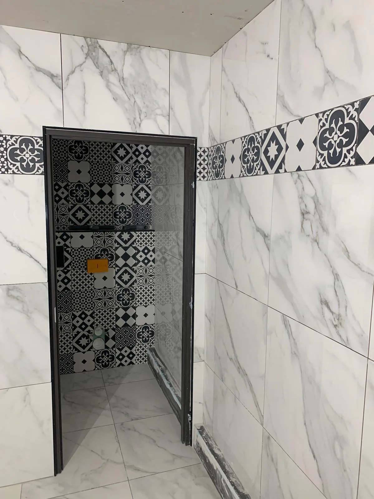 Cabine de douche vue d'angle, murs en faïence grand format effet marbre blanc, frise horizontale et fond en carreaux à motifs noir et blanc, paroi vitrée à profilé sombre