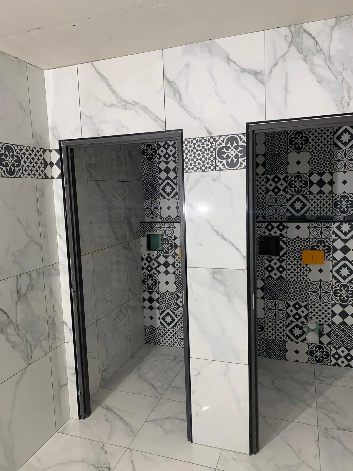 Deux cabines de douche côte à côte aux murs en faïence grand format effet marbre blanc, fond et frise en carreaux à motifs noir et blanc, niches intégrées