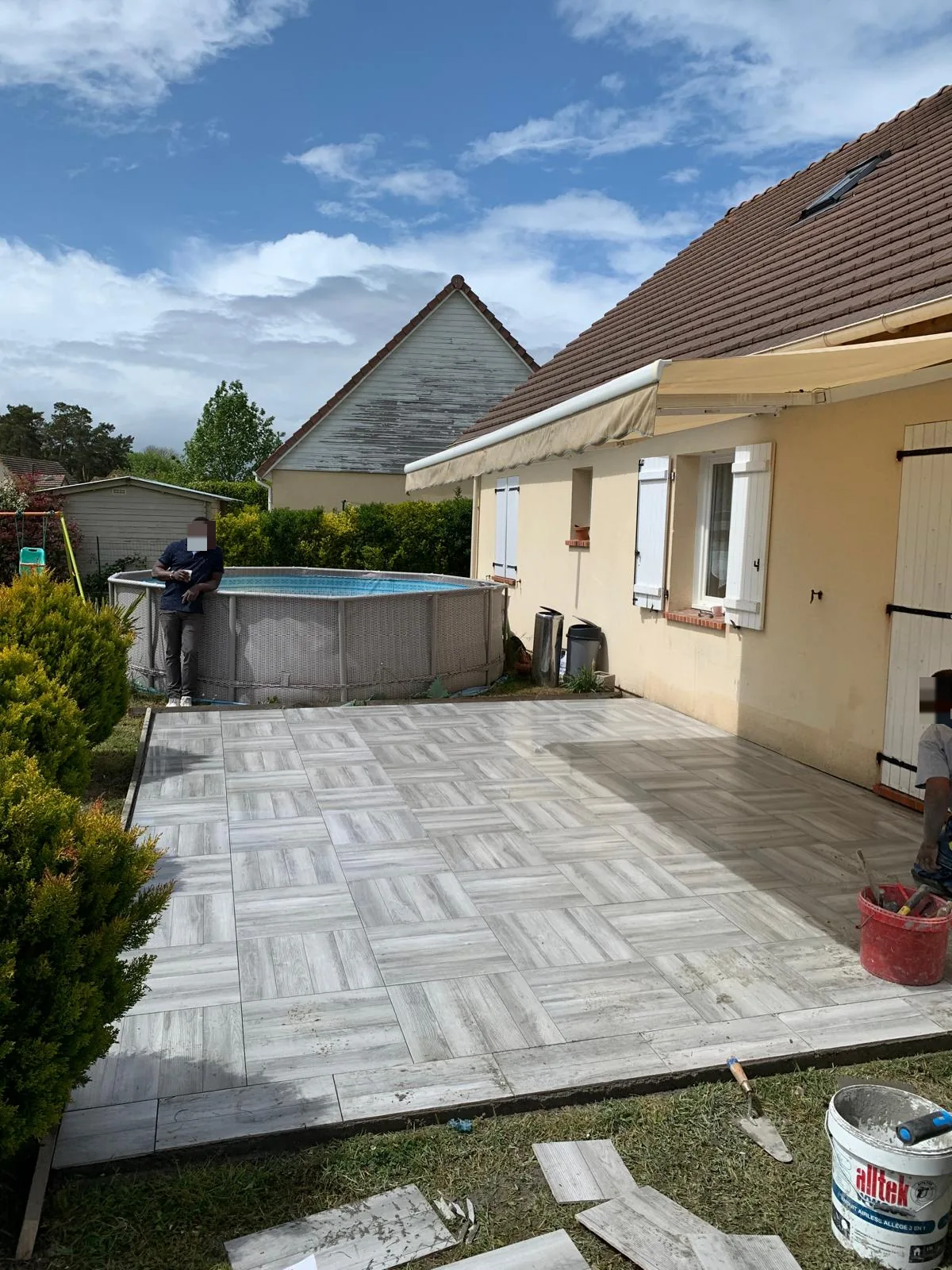 Terrasse extérieure en carrelage imitation bois clair posé en motif damier, le long d'une maison, avec une piscine hors-sol et un jardin en arrière-plan