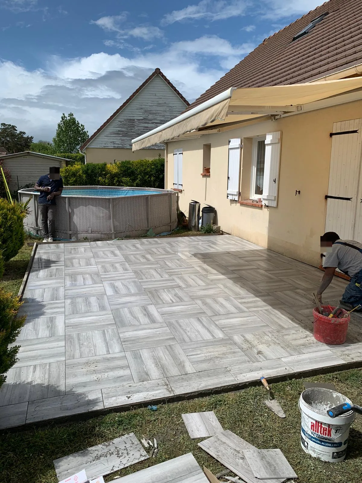 Terrasse exterieure en carrelage imitation bois clair pose en damier de lames devant une maison, avec une piscine hors-sol a gauche