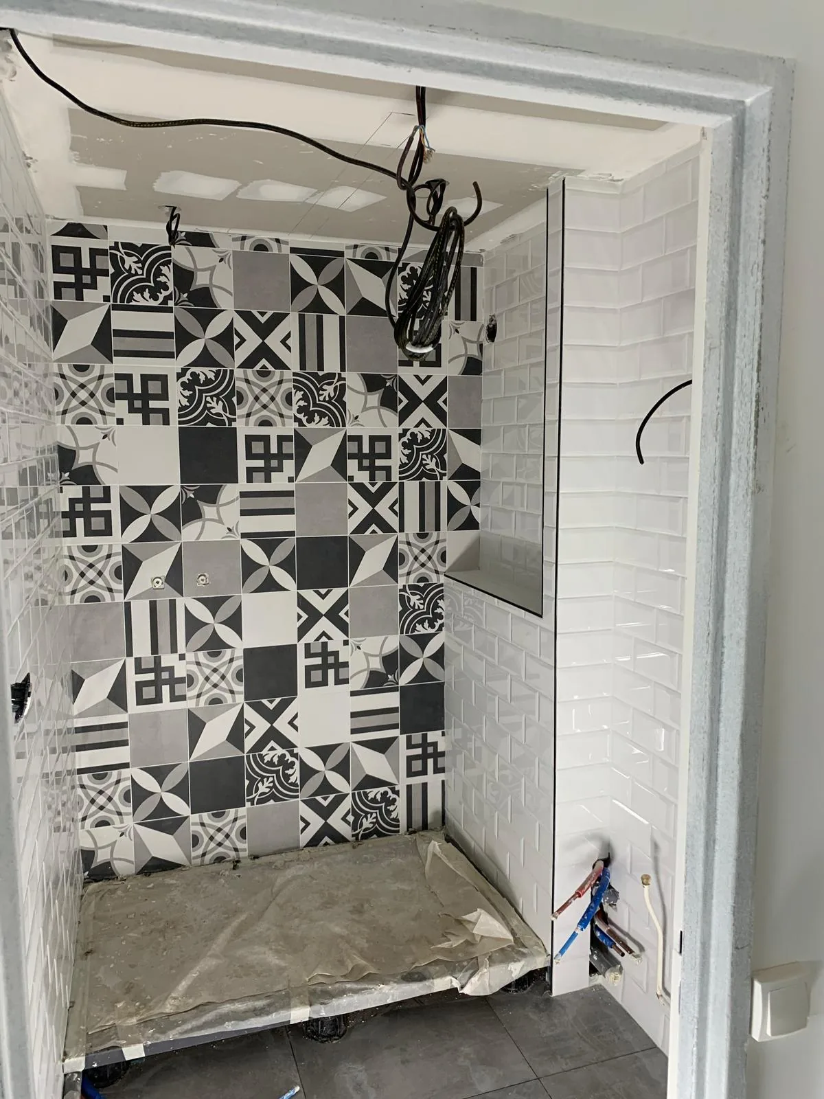 Niche de douche carrelee de carreaux type ciment a motifs geometriques noir, blanc et gris, avec un mur lateral en faience metro blanche biseautee et une tablette integree