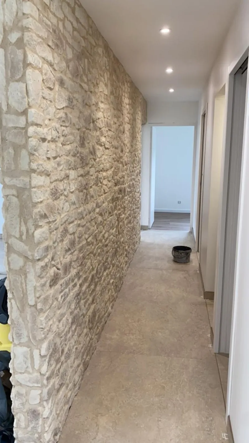 Couloir habille d un mur en pierre de parement beige, sol carrele en grand format beige et spots encastres au plafond