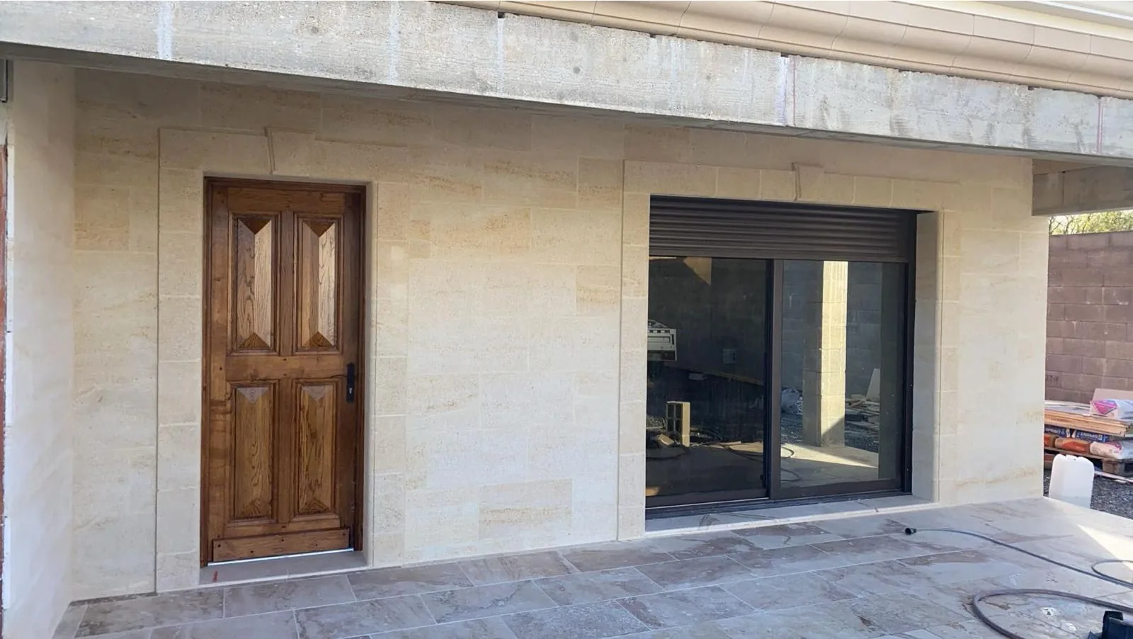 Facade exterieure habillee de plaquettes de parement aspect pierre de taille en appareil regulier, encadrant une porte en bois massif et une grande baie coulissante a chassis noir ; sol de terrasse en carrelage aspect pierre claire