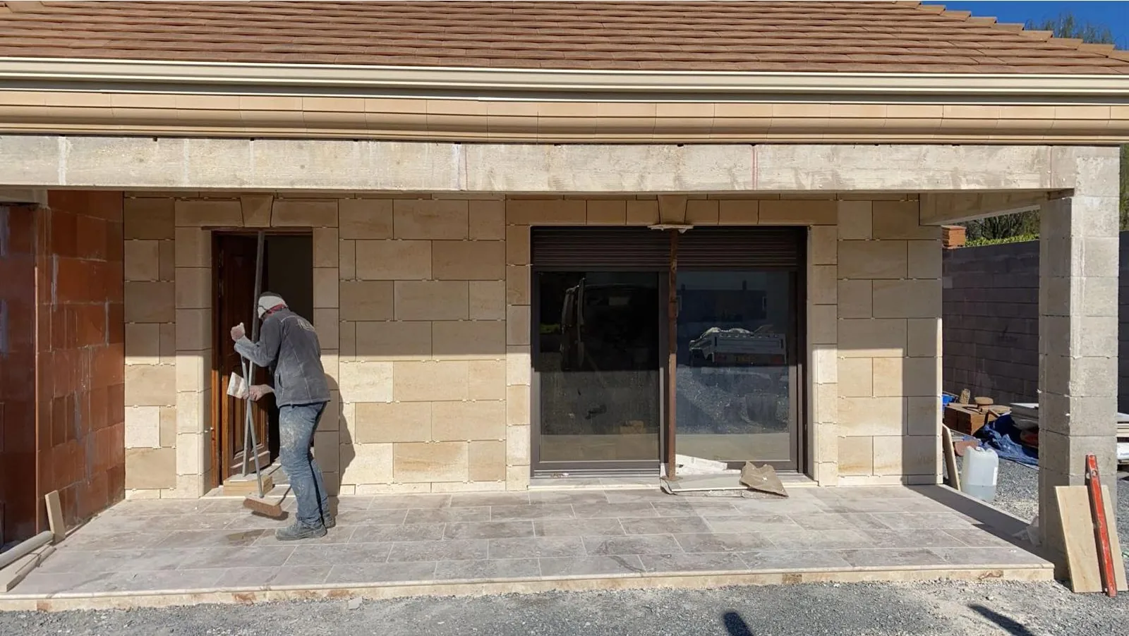 Ouvrier vu de dos, sur une planche d echafaudage, posant des plaquettes de parement aspect pierre sur une facade autour d une porte en bois massif
