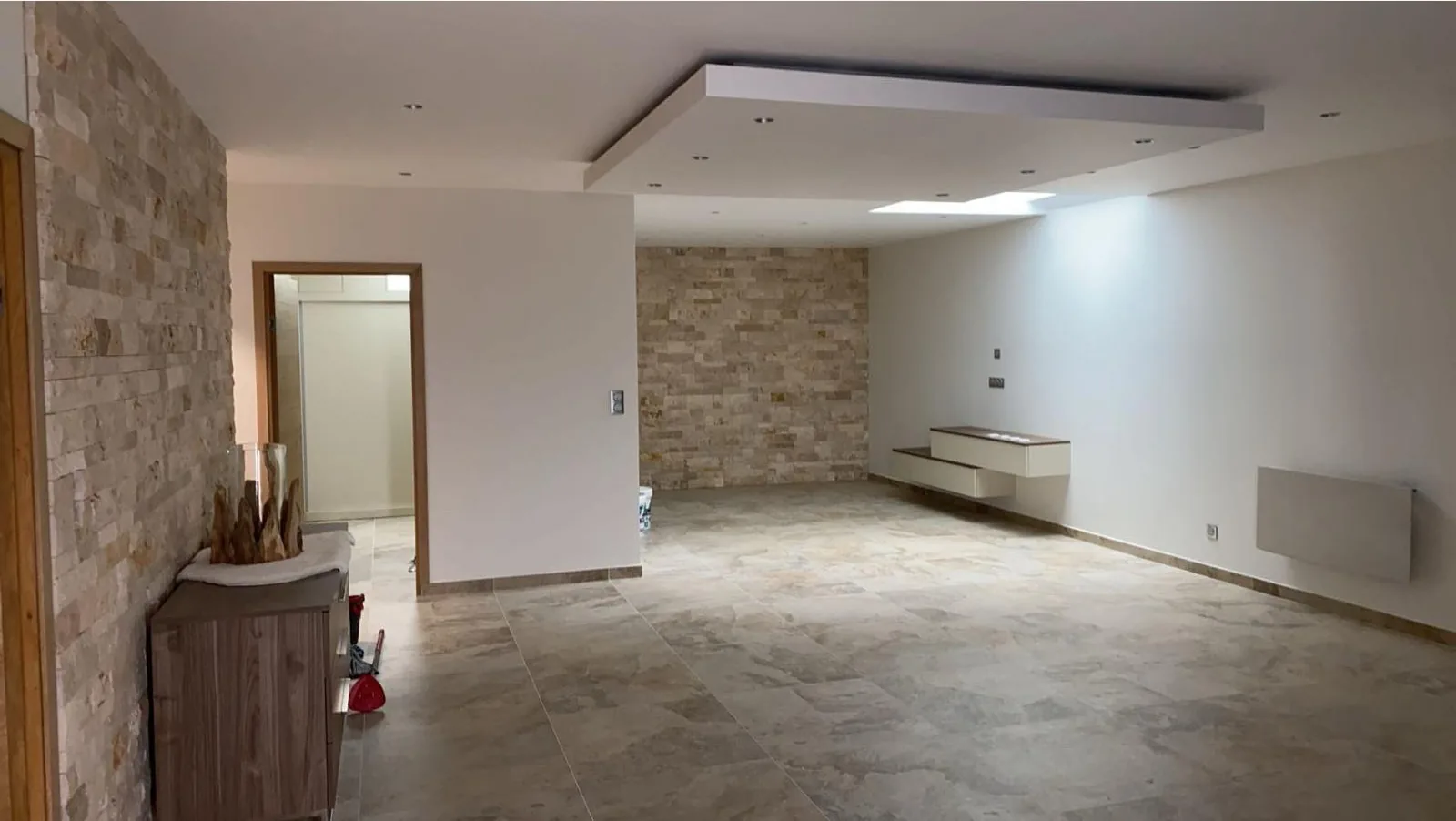 Sejour termine avec un sol en grands carreaux aspect travertin beige, un mur d accent habille de plaquettes de parement pierre, un faux plafond a eclairage encastre et un buffet en bois