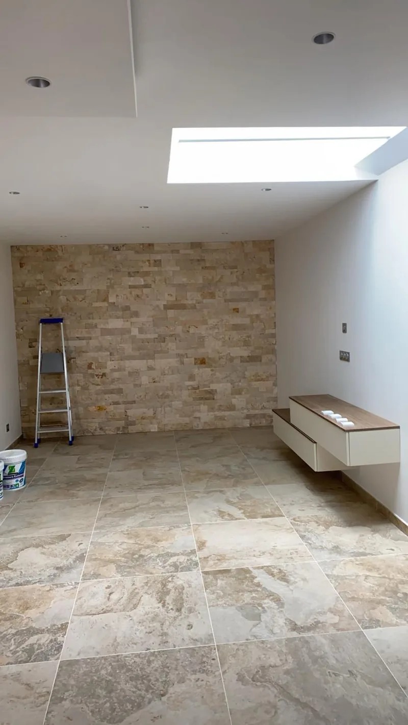 Piece a vivre au sol carrele de grands carreaux carres aspect travertin tres veine, avec un mur d accent en plaquettes de parement pierre, un faux plafond a puits de lumiere et un meuble suspendu a plateau bois