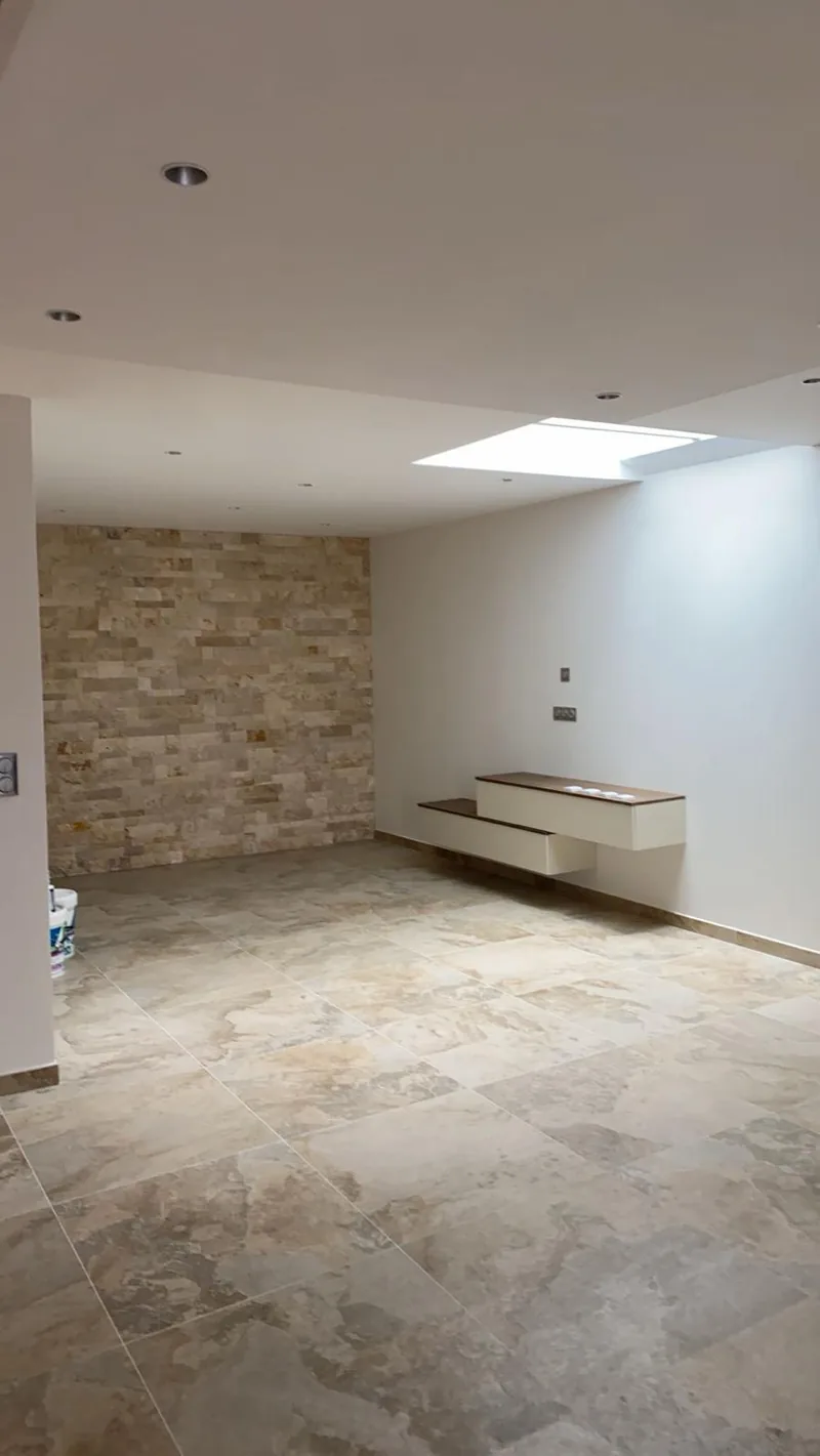 Piece a vivre vide au sol carrele de grands carreaux carres aspect travertin, avec un mur d accent en plaquettes de parement pierre, un puits de lumiere zenithal et un meuble suspendu a plateau bois