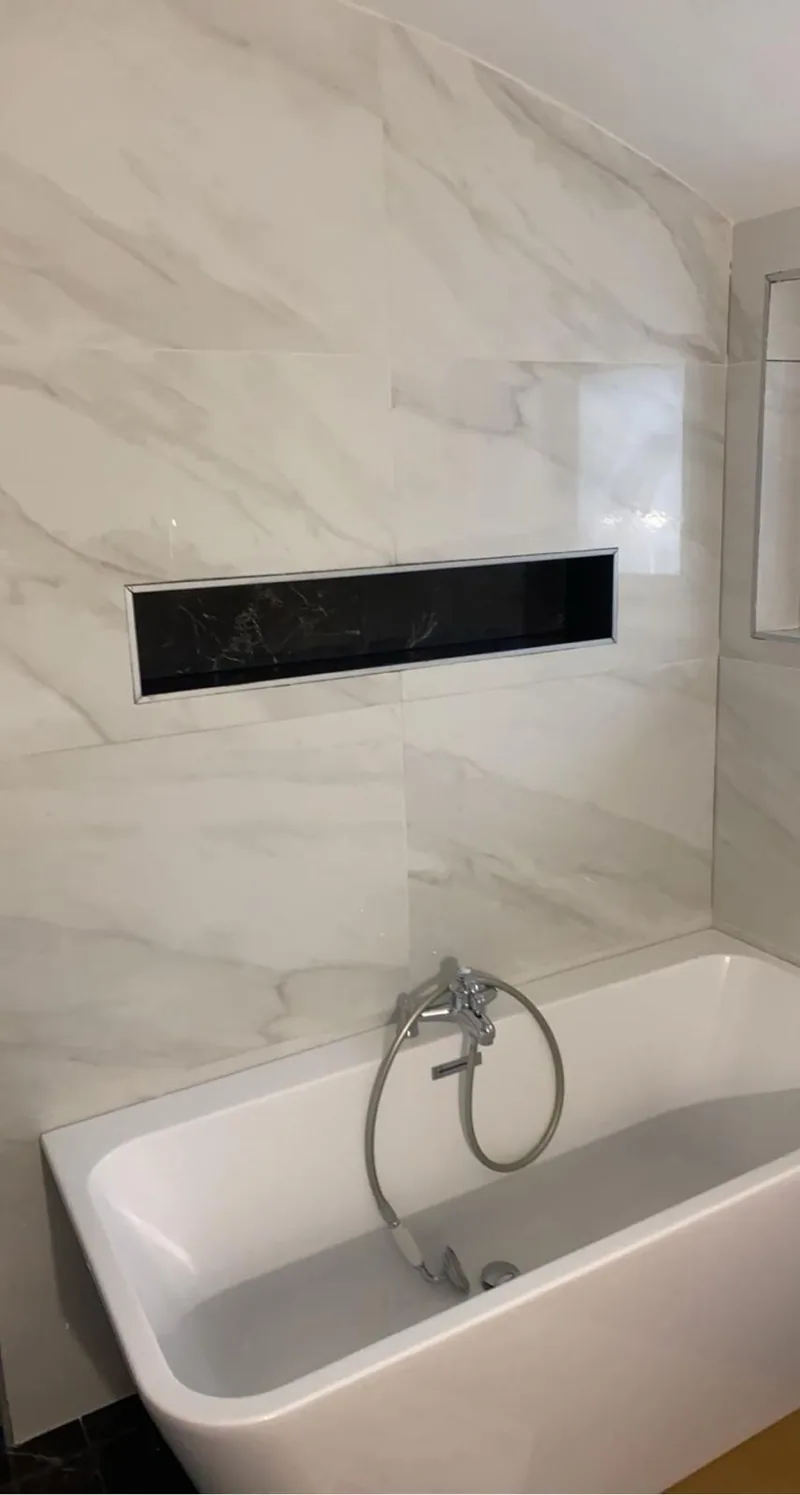 Mur de salle de bains en faience grand format effet marbre blanc veine de gris, brillante, avec une longue niche horizontale encastree a fond effet marbre noir, au-dessus d une baignoire equipee d un mitigeur et d une douchette chromes