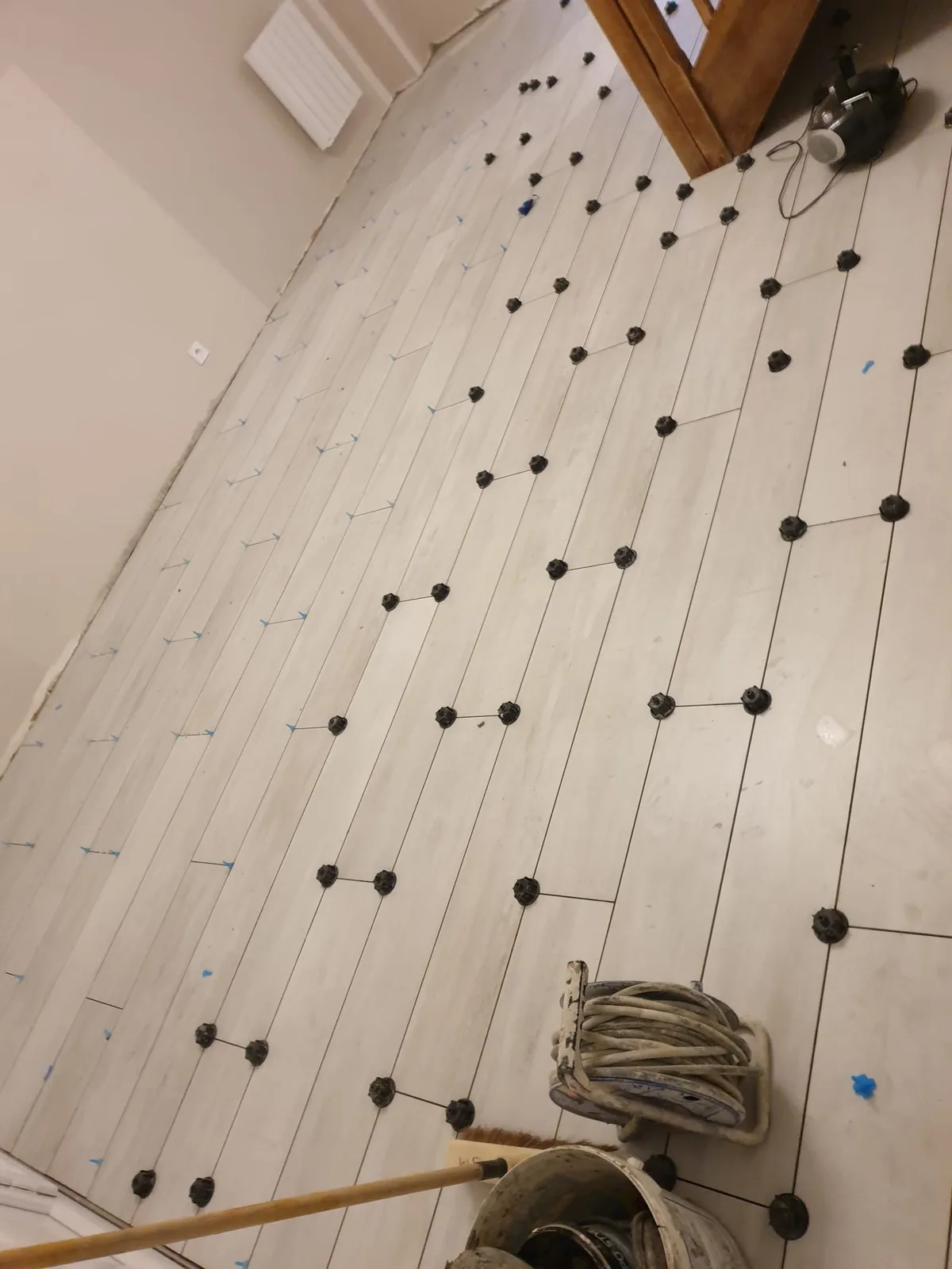 Sol en cours de carrelage avec des lames claires veinées posées à joints décalés, croisillons autonivelants noirs répartis sur les joints, dévidoir et seau de chantier au fond