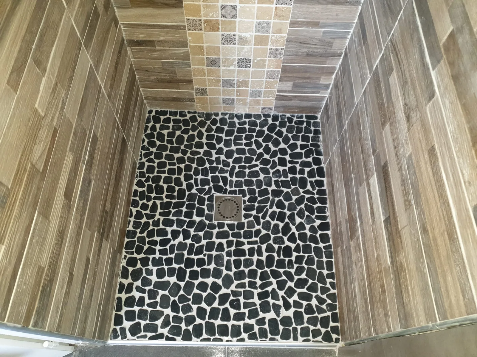Douche à l'italienne vue de dessus, sol en galets noirs jointoyés de blanc autour d'un siphon carré, murs en faïence imitation bois et bandeau vertical de mosaïque beige à motifs façon carreaux de ciment