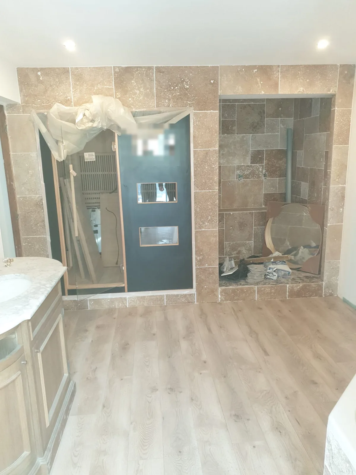 Salle de bains en rénovation avec habillage mural en carrelage aspect travertin, niche de douche carrelée à droite, portes vitrées à gauche et sol aspect bois, meuble vasque ancien au premier plan