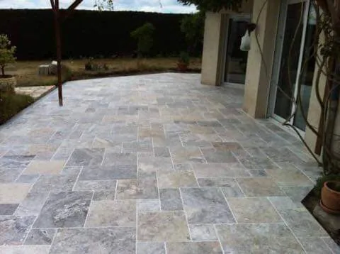 Terrasse extérieure pavée en dalles aspect travertin gris posées en opus romain, bordée d'une façade claire avec baie coulissante à droite et d'un jardin avec haie au fond