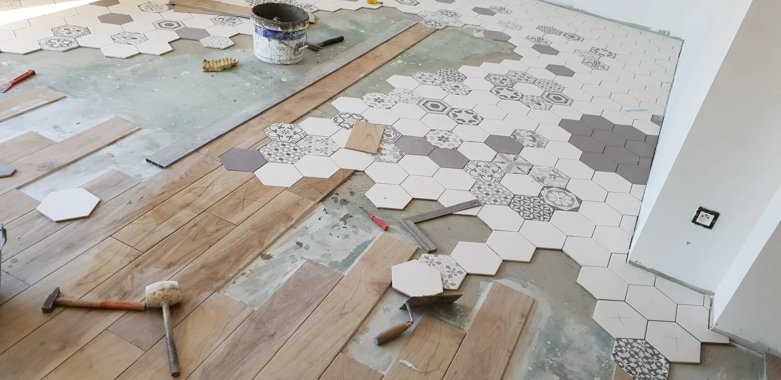 Sol en cours de pose associant des carreaux hexagonaux blancs, gris et à motifs de carreaux de ciment à un carrelage imitation bois, avec maillet, équerre et seau de colle posés au sol