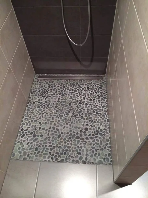 Douche à l'italienne terminée, sol en galets gris jointoyés avec caniveau linéaire inox au fond, murs en carrelage grand format taupe et paroi du fond anthracite