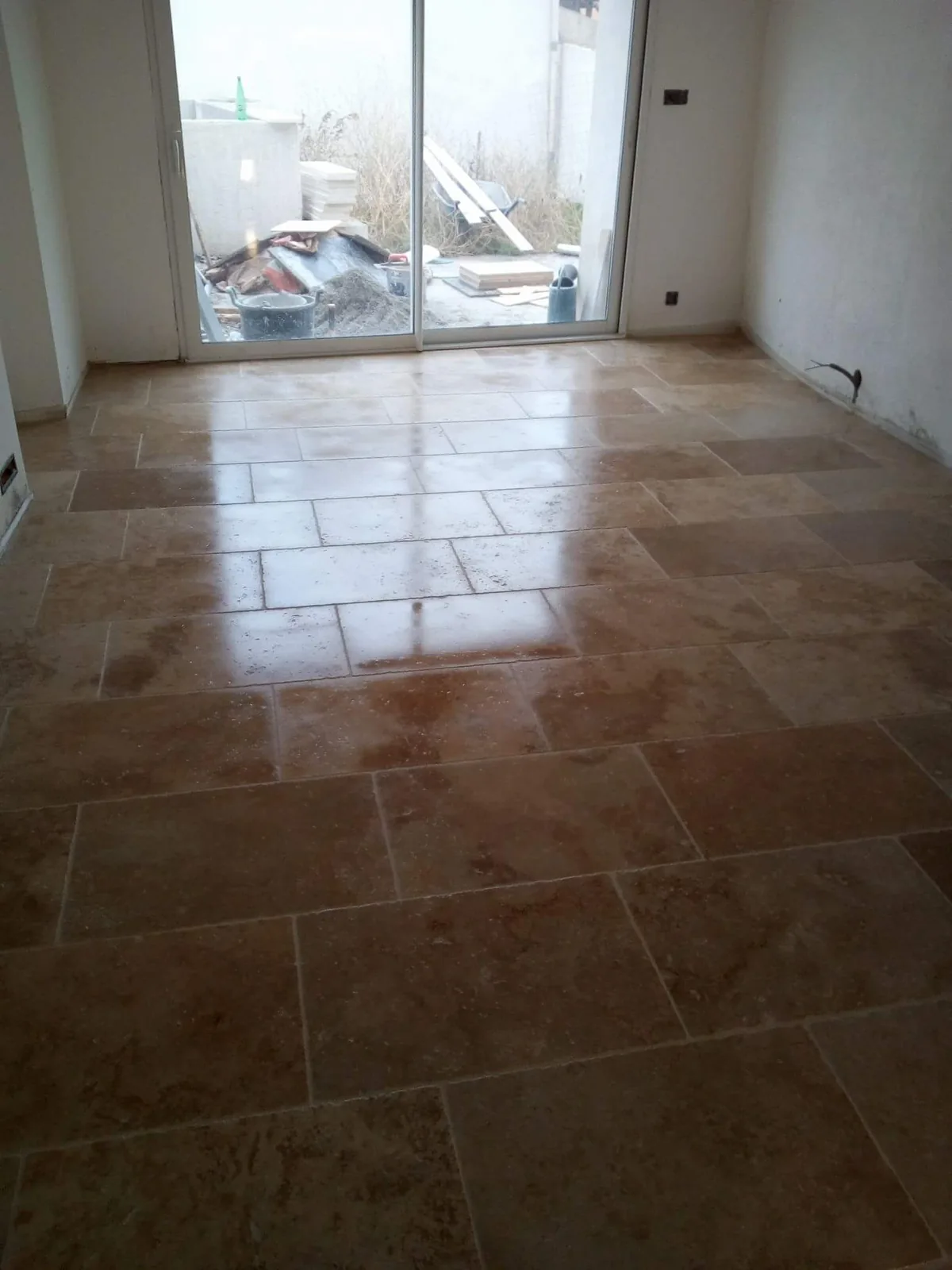 Pièce vide au sol carrelé en aspect travertin beige posé en opus romain, encore humide après jointoiement, baie coulissante ouvrant sur un chantier au fond