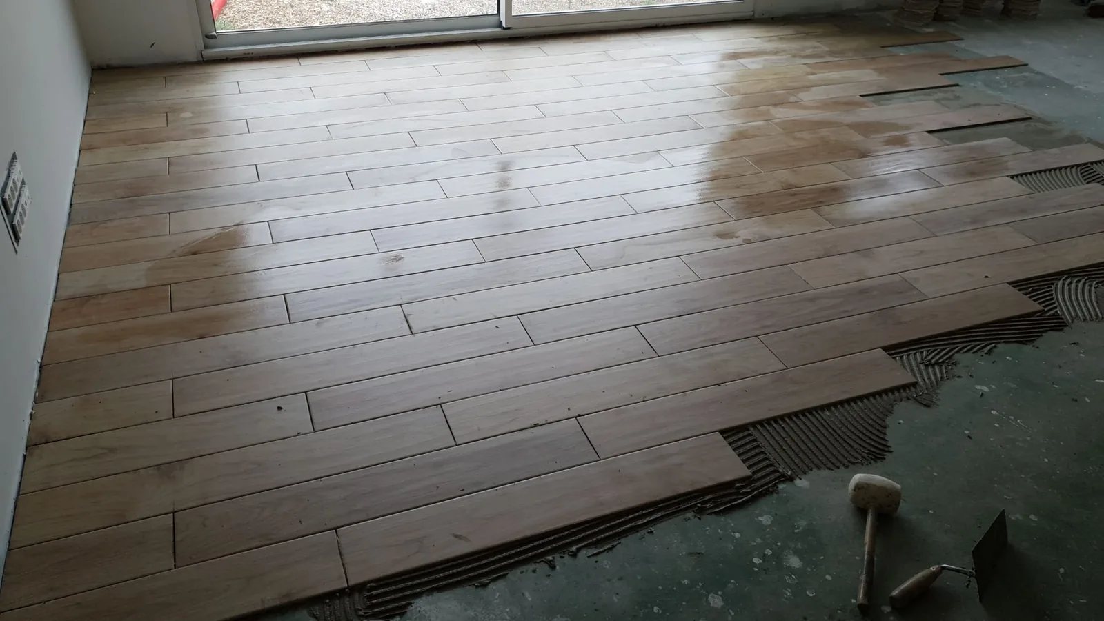 Sol en cours de carrelage avec des lames imitation bois clair posées à joints décalés, colle peignée visible sur la chape à droite, maillet et truelle posés au sol