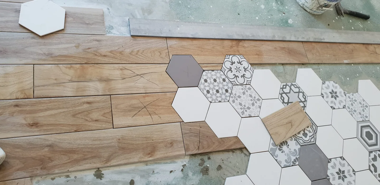 Vue rapprochee en plongee d'un sol en cours de pose : des lames de carrelage imitation bois s'imbriquent avec des carreaux hexagonaux blancs, gris et a motifs. Des reperes au crayon marquent les carreaux a couper.